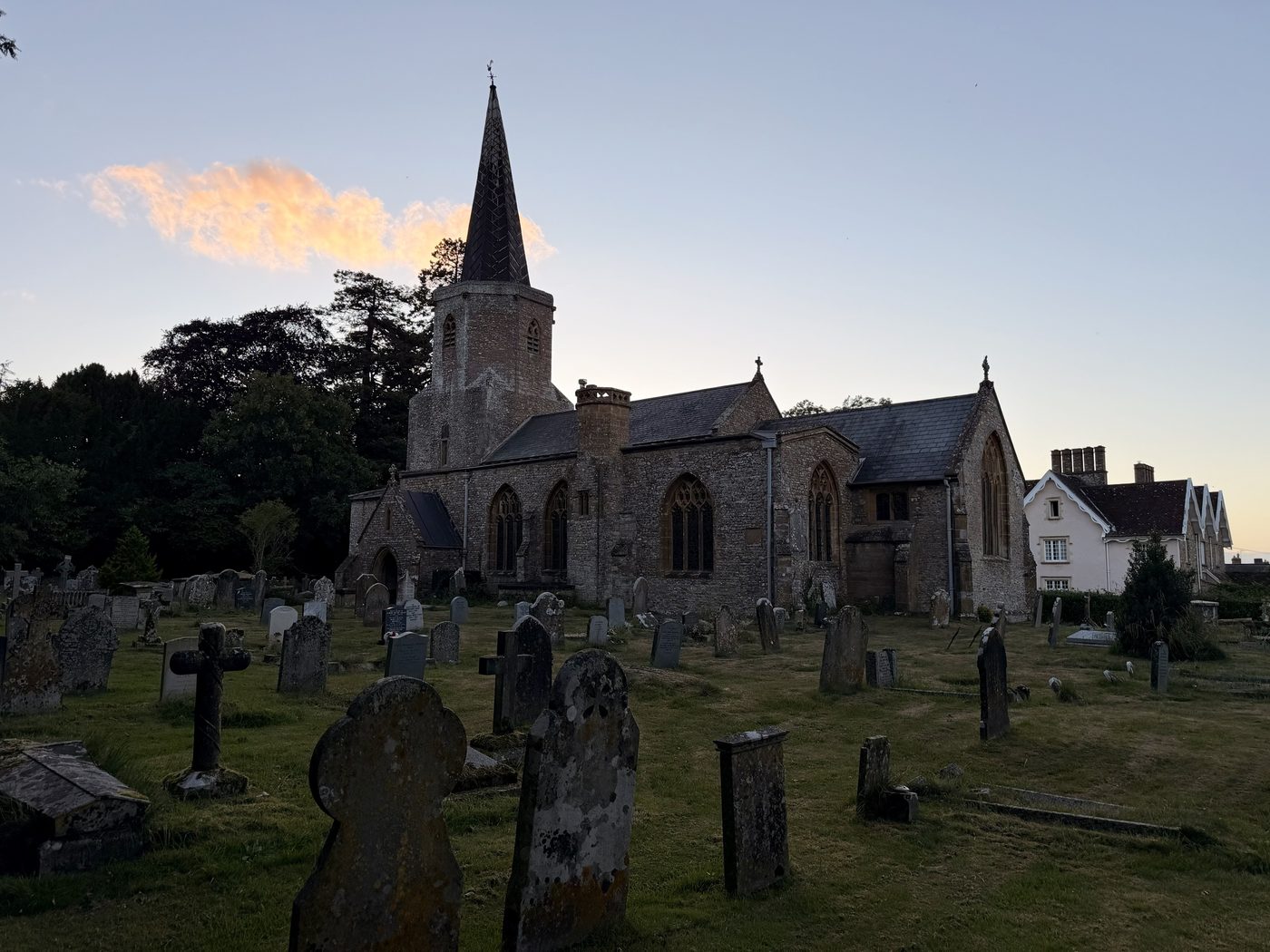 The church of St Andrew and St Mary, Pitminster, at dusk, seen across the churchyard: the tower rising to its octagon and lead spire, a lit cloud behind